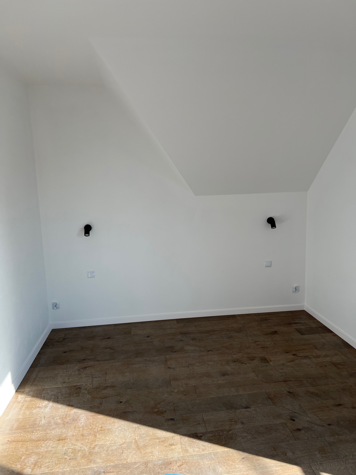 Pusta, jasna przestrzeń pod skosem z drewnianą podłogą, białymi ścianami, dwoma czarnymi kinkietami i gniazdkami elektrycznymi, oświetlona naturalnym światłem.