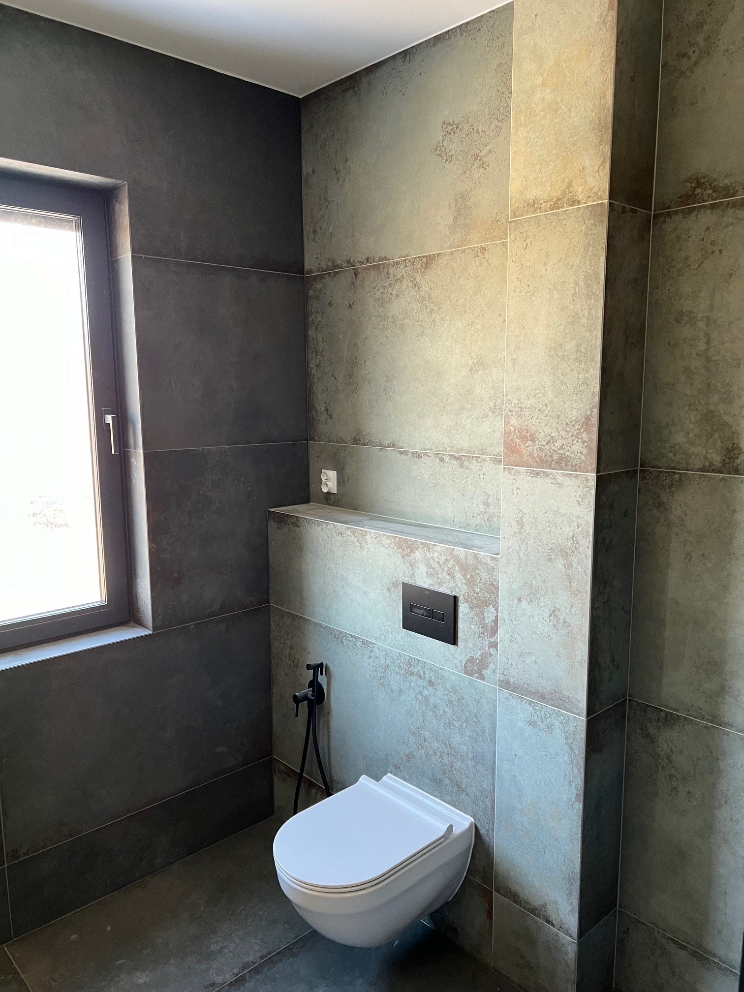Nowoczesna łazienka z toaletą podwieszaną, bidetką w kolorze czarnym i płytkami imitującymi kamień w odcieniach szarości i beżu, oświetlona naturalnym światłem z okna.