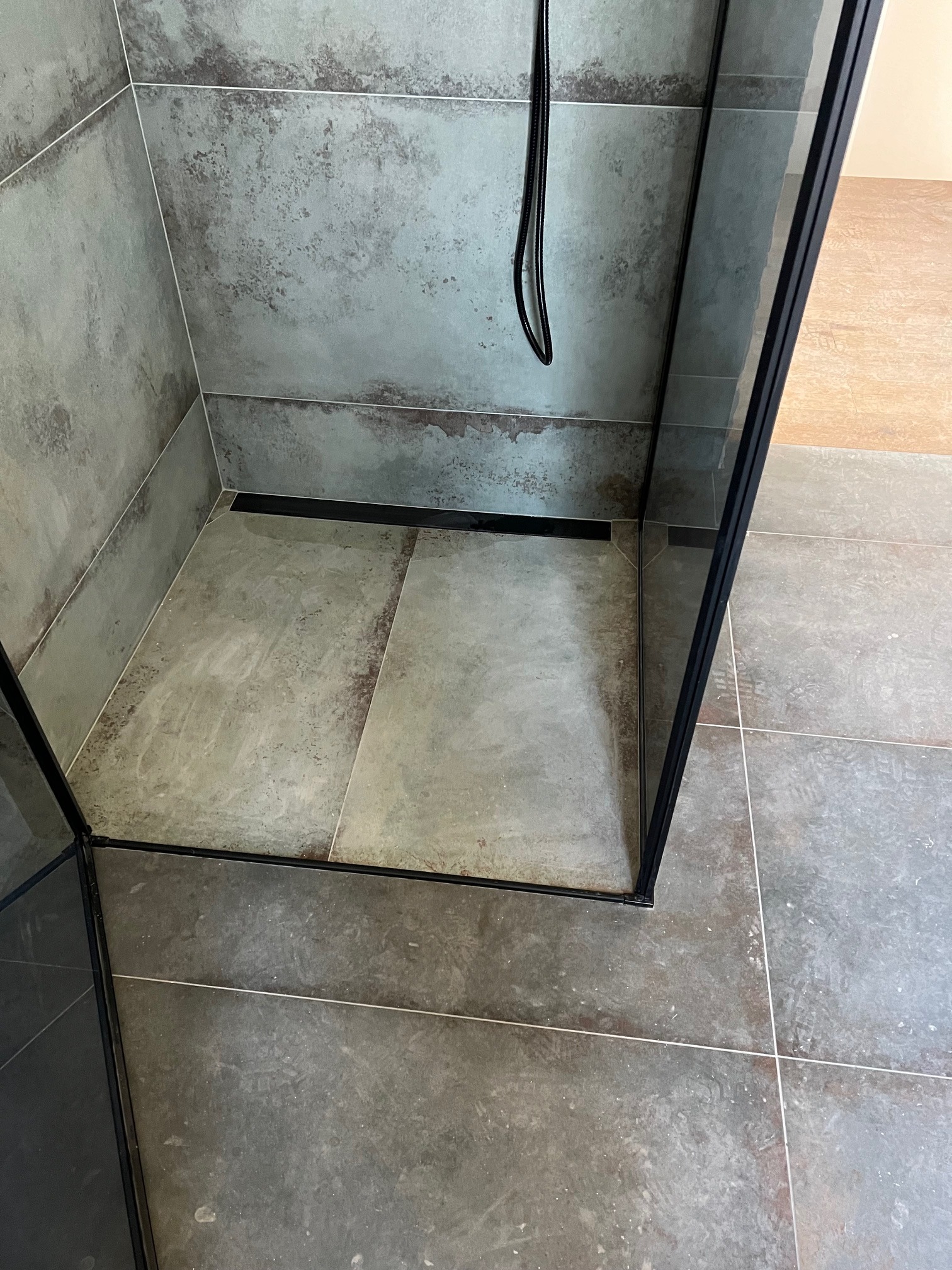 Nowoczesna kabina prysznicowa typu walk-in z szarymi płytkami imitującymi beton, czarnym odpływem liniowym i czarną ramą kabiny