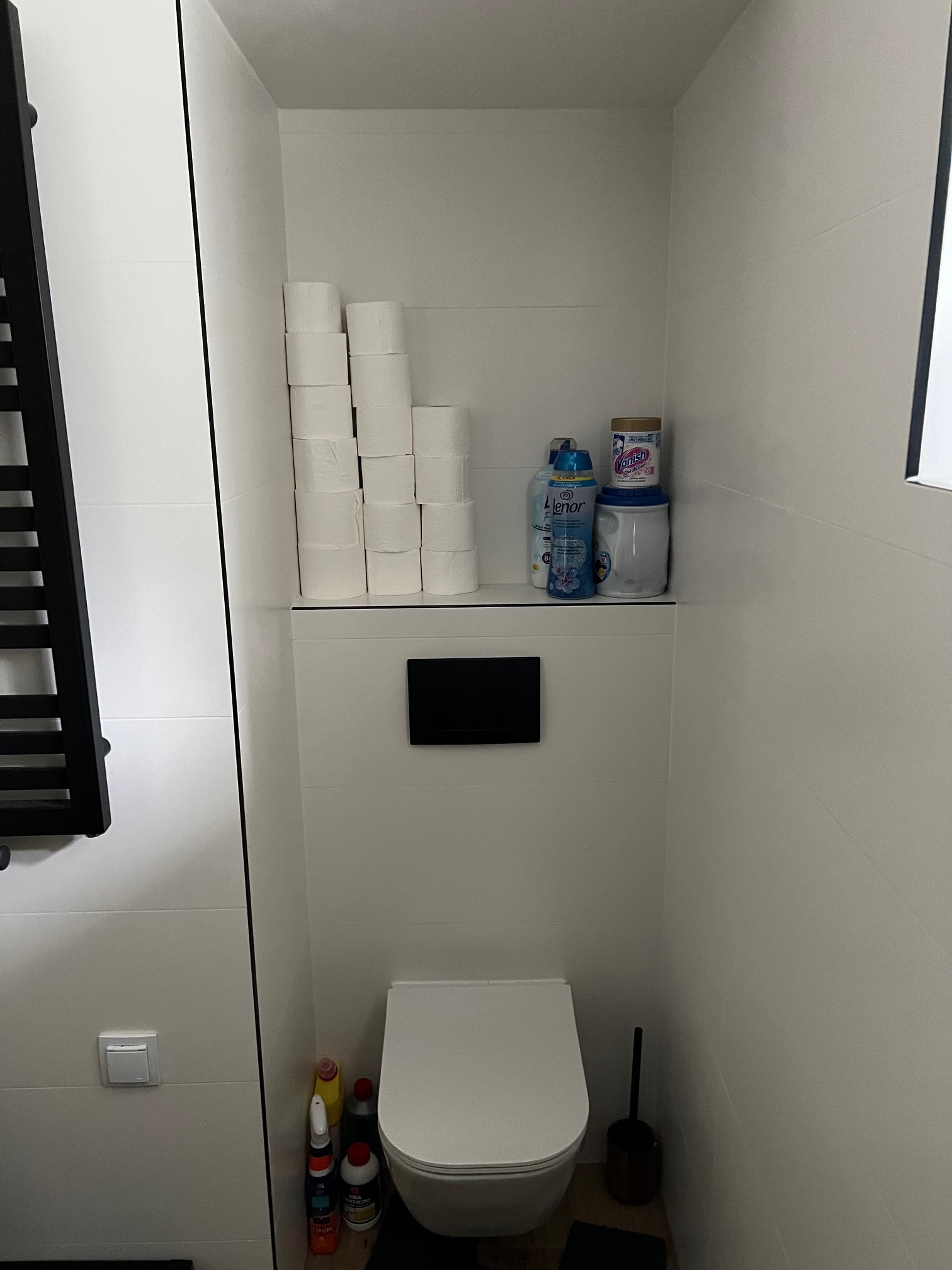 Biała, minimalistyczna toaleta z podwieszaną muszlą, czarnym grzejnikiem drabinkowym i półką z zapasem papieru oraz środkami czystości. Ściany wyłożone białymi płytkami.