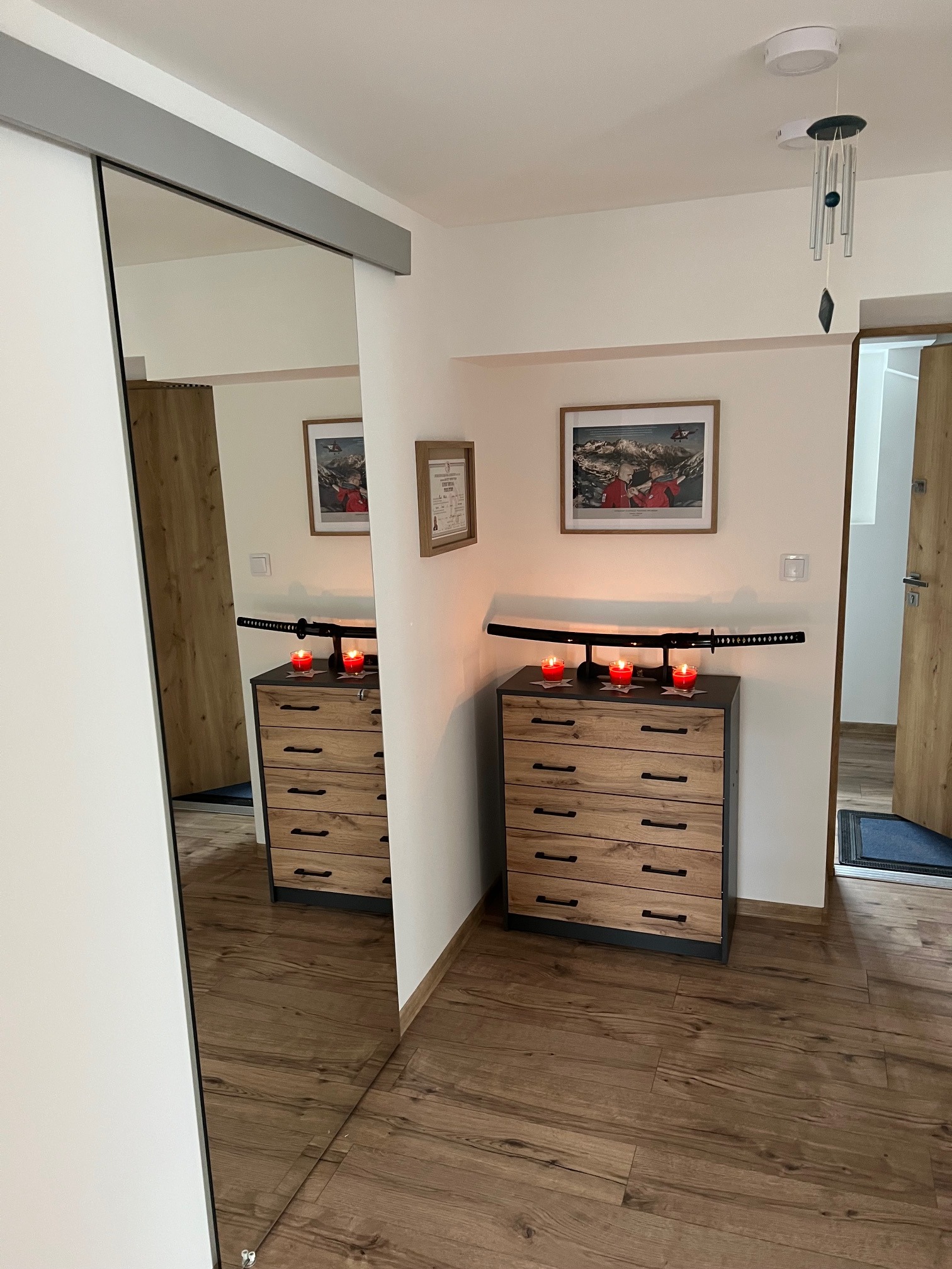 Nowoczesna komoda z drewnianymi frontami szuflad, ozdobiona kataną i świecami. Widok z perspektywy lustra, ukazujący fragment wnętrza z drewnianą podłogą.