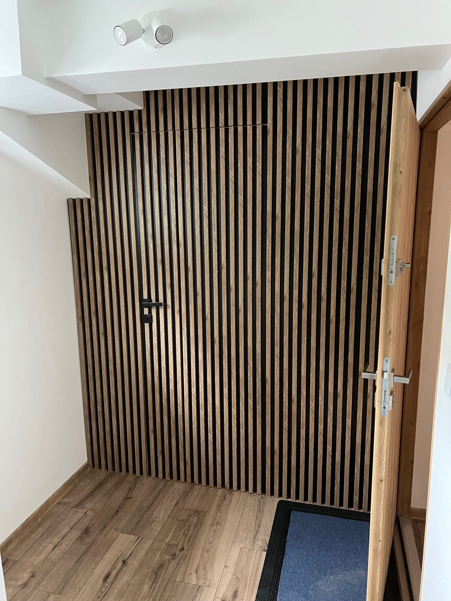 Drewniane lamele na ścianie korytarza, ukrywające drzwi, z otwartymi drzwiami po prawej stronie i fragmentem drewnianej podłogi. Nowoczesny design.