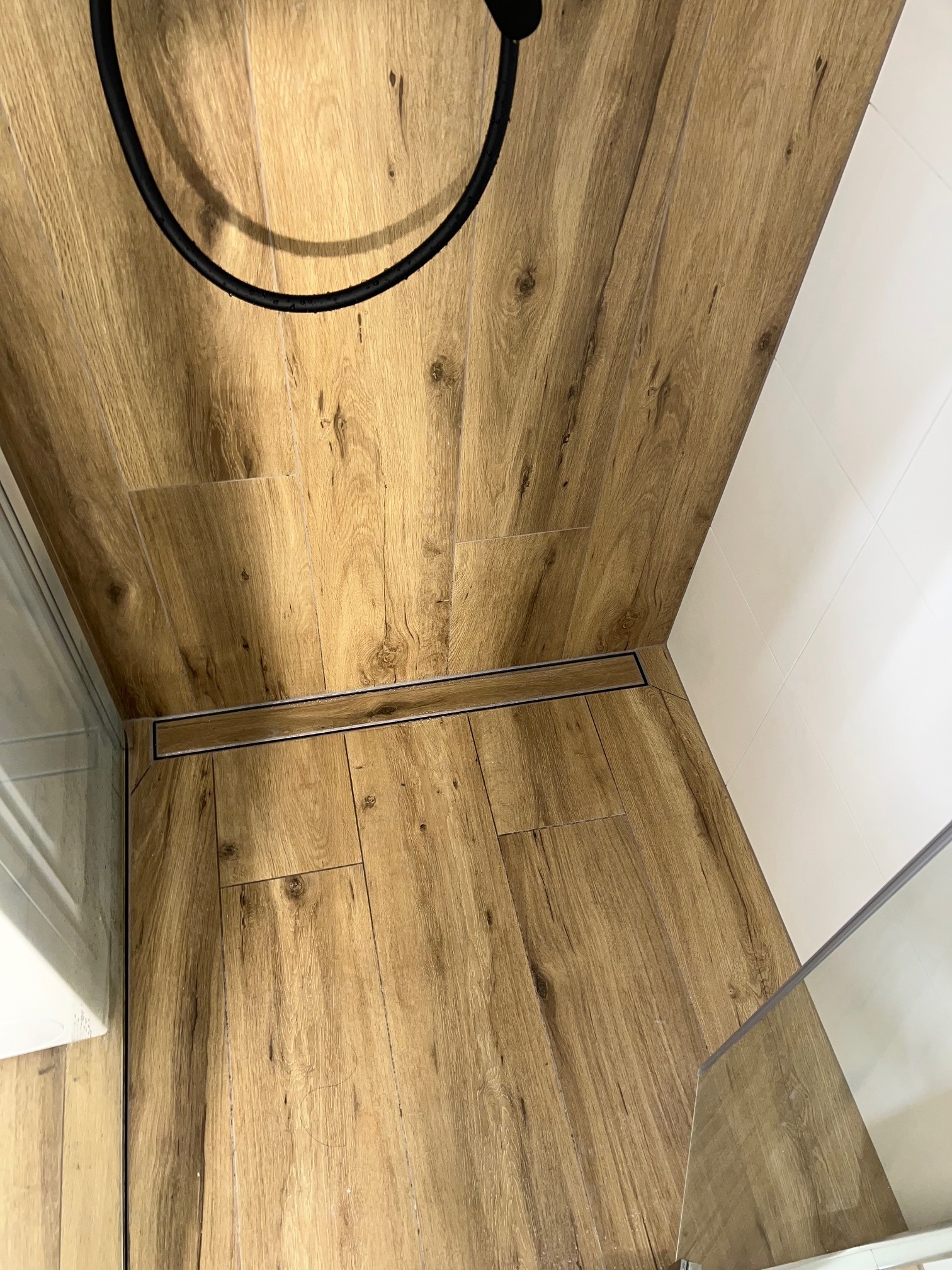 Parkiet w łazience z odpływem liniowym, kabina prysznicowa ze szklaną ścianką, białe płytki na ścianie. Nowoczesne wykończenie wnętrza łazienki.