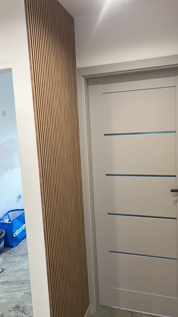 Panel lamelowy na ścianie obok białych drzwi z poziomymi wstawkami. Widoczny fragment pomieszczenia w trakcie prac wykończeniowych.