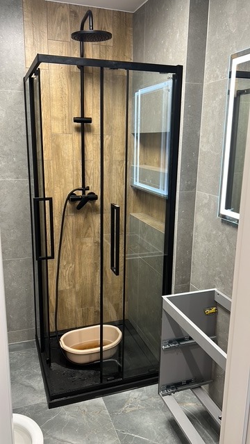 Nowoczesna kabina prysznicowa z czarnymi profilami i deszczownicą, drewnopodobne płytki wewnątrz, szare płytki na ścianach, niedokończona szafka łazienkowa obok.