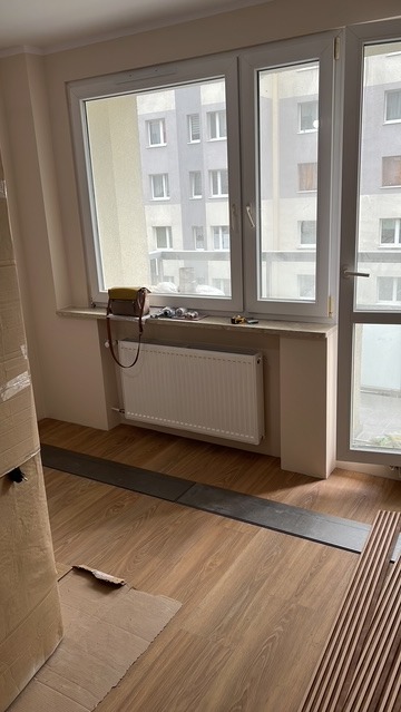 Montaż paneli podłogowych w jasnym pokoju z grzejnikiem pod oknem. Widoczne narzędzia i materiały, częściowo zakryte kartonem. Ujęcie z niskiego kąta.