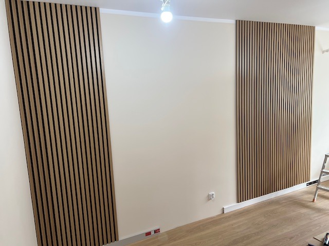 Ściana z drewnianymi lamelami w jasnym wnętrzu. Dwie pionowe sekcje lameli po bokach ściany, widoczne gniazdko elektryczne i fragment drabiny.