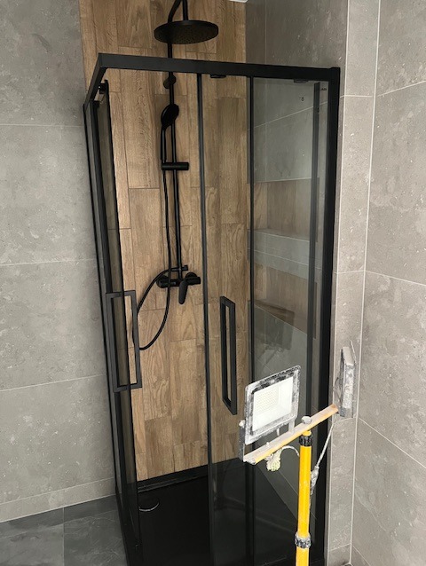 Kabina prysznicowa z czarnymi profilami i deszczownicą, wykończona w Sosnowcu. Widoczne płytki imitujące drewno i szara ściana. Oświetlenie robocze.