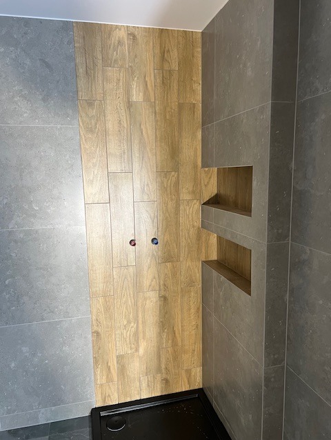 Wykończona kabina prysznicowa z drewnopodobnymi płytkami i wnękami na kosmetyki, szare płytki na pozostałych ścianach. Widoczne przyłącza wodne.