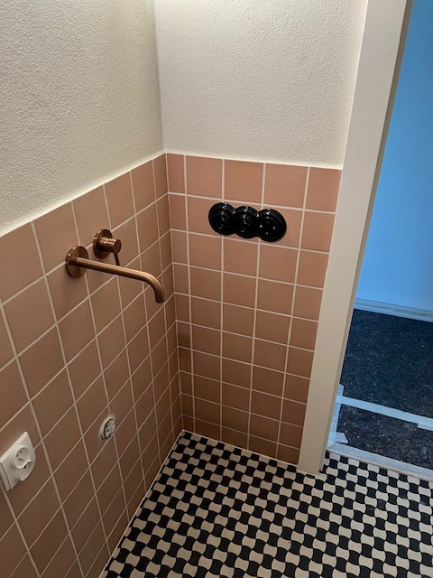 Łazienka po renowacji: różowe płytki ścienne, miedziana bateria, czarno-biała podłoga w szachownicę i czarne włączniki. Nowoczesny design z nutą retro.