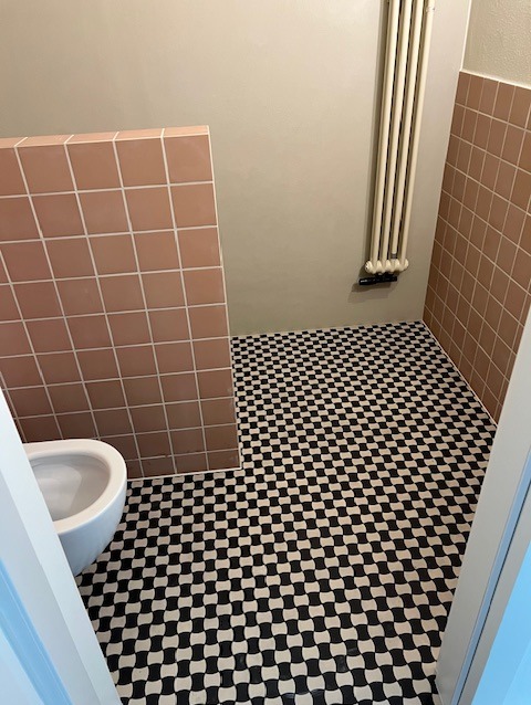 Wykończona łazienka z czarno-białą szachownicą na podłodze i różowymi płytkami na ścianie. Grzejnik żeberkowy na ścianie. Widok zza framugi drzwi.