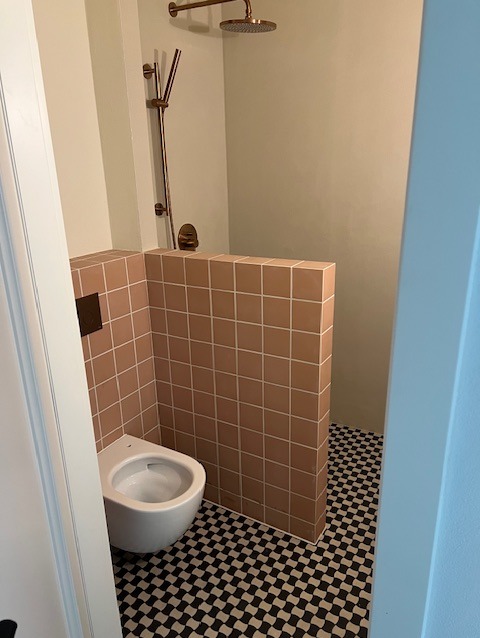 Nowoczesna łazienka z kabiną prysznicową oddzieloną niską ścianką z beżowych płytek. Umywalka podwieszana, podłoga w czarno-białą szachownicę, armatura w kolorze miedzi.