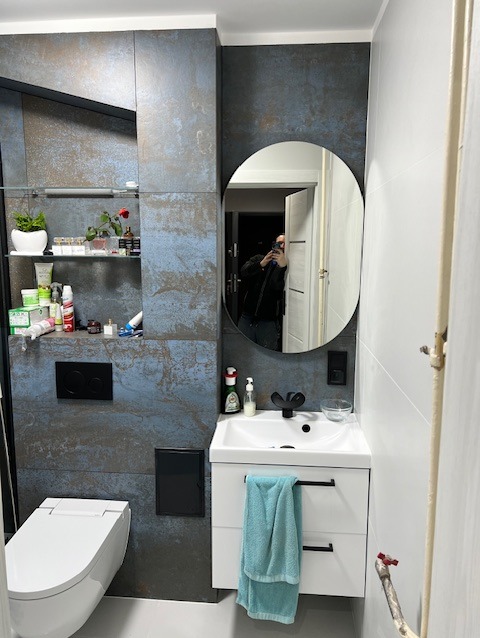 Nowoczesna łazienka z płytkami imitującymi rdzę, owalnym lustrem i białą szafką umywalkową z turkusowym ręcznikiem. Widoczna toaleta i półki z kosmetykami.