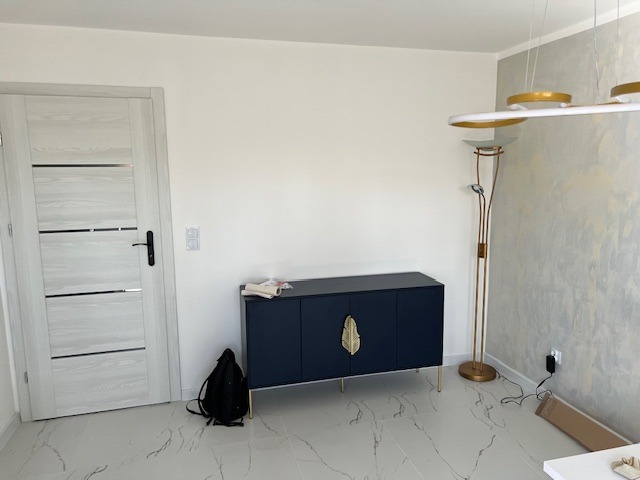 Nowoczesne wnętrze: granatowa komoda z złotymi uchwytami, designerska lampa podłogowa i drzwi z poziomymi przeszkleniami. Minimalistyczny styl, jasne ściany i marmurowa podłoga.