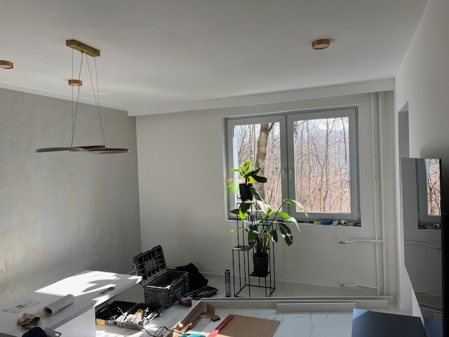 Wnętrze pokoju w Sosnowcu w trakcie wykańczania. Widoczne narzędzia, roślina doniczkowa przy oknie i nowoczesna lampa wisząca. Ściana częściowo pomalowana.
