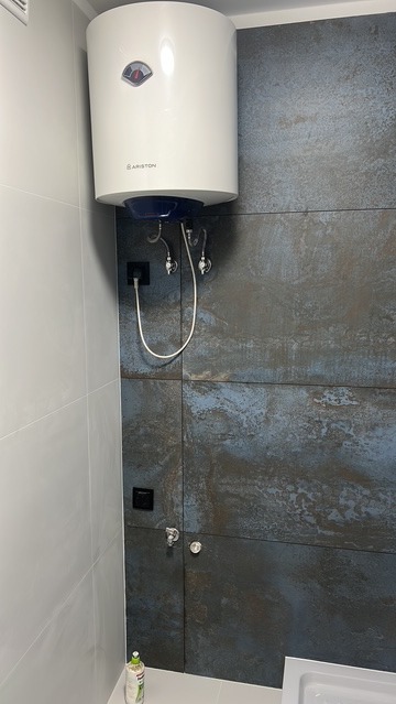 Bojler Ariston zamontowany w łazience z szaro-niebieskimi płytkami imitującymi metal. Widoczne przyłącza wodne i elektryczne. Minimalistyczny design.