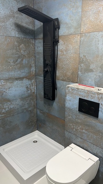 Nowoczesna kabina prysznicowa z panelem natryskowym w kolorze czarnym, biały brodzik i toaleta. Ściany wyłożone płytkami imitującymi kamień.