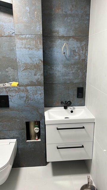 Nowoczesna łazienka z umywalką na szafce, armaturą w kolorze czarnym i płytkami imitującymi rdzę. Widoczny fragment toalety i instalacja wodna w ścianie.