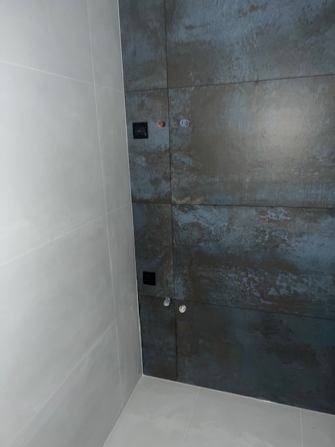 Wykończona łazienka z szarymi, matowymi płytkami na ścianach i podłodze. Na jednej ze ścian widoczne przyłącza wodne i gniazdko elektryczne.