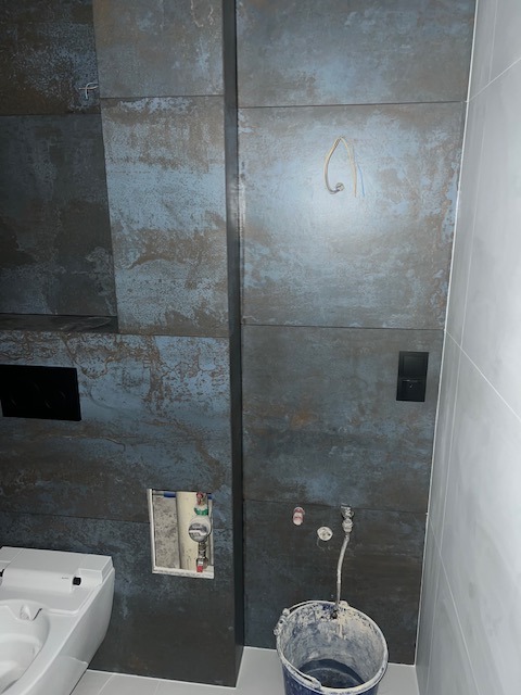 Łazienka w trakcie remontu: położone płytki imitujące rdzę, widoczne przyłącza wodne, otwór rewizyjny, częściowo widoczna toaleta i wiadro z zaprawą.