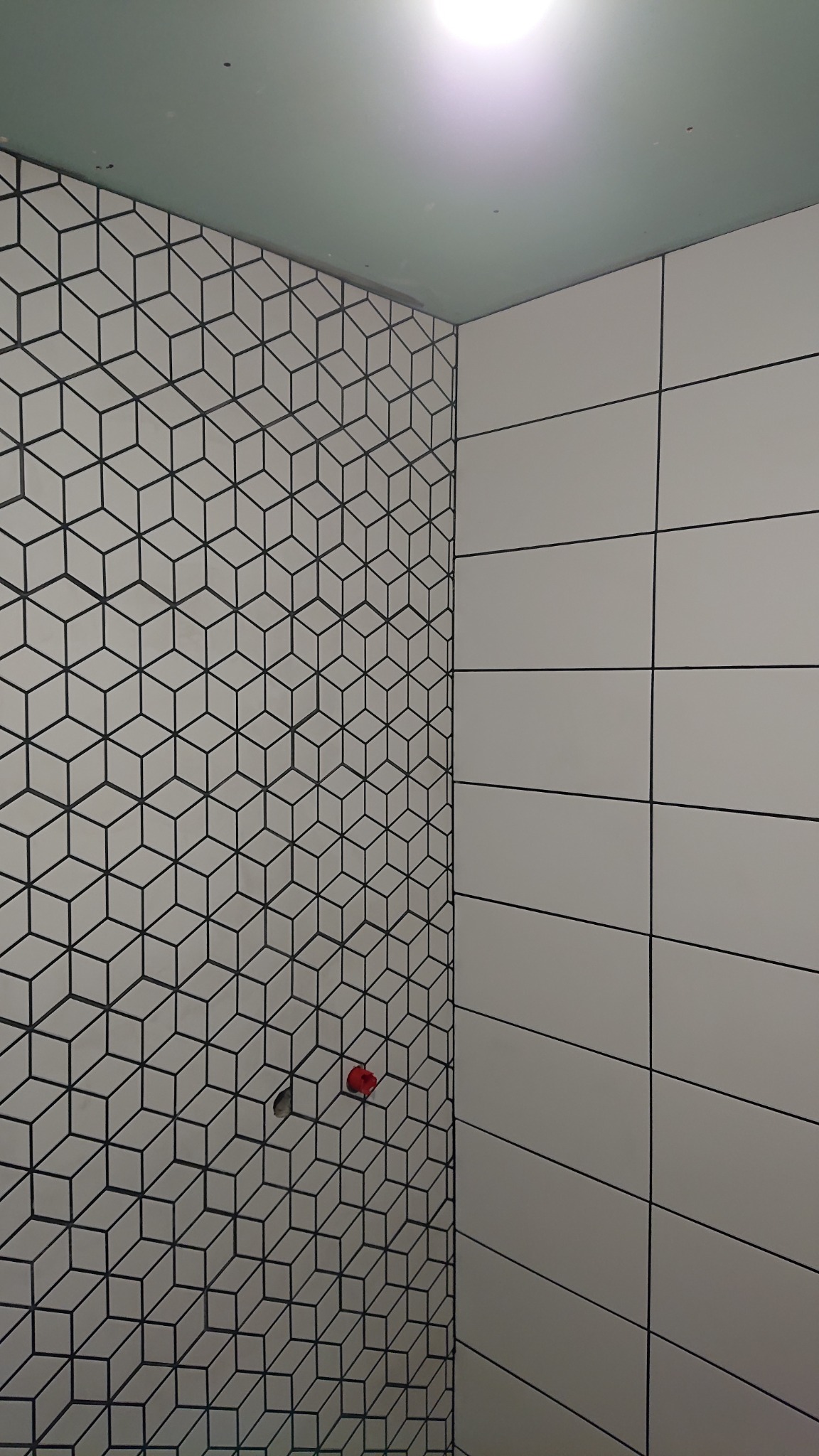 Ściana łazienki wyłożona białymi płytkami z czarnym wzorem geometrycznym tworzącym iluzję kostek 3D, sąsiadująca ze ścianą z prostokątnych, białych płytek z czarnymi fugami, widoczny czerwony zawór...