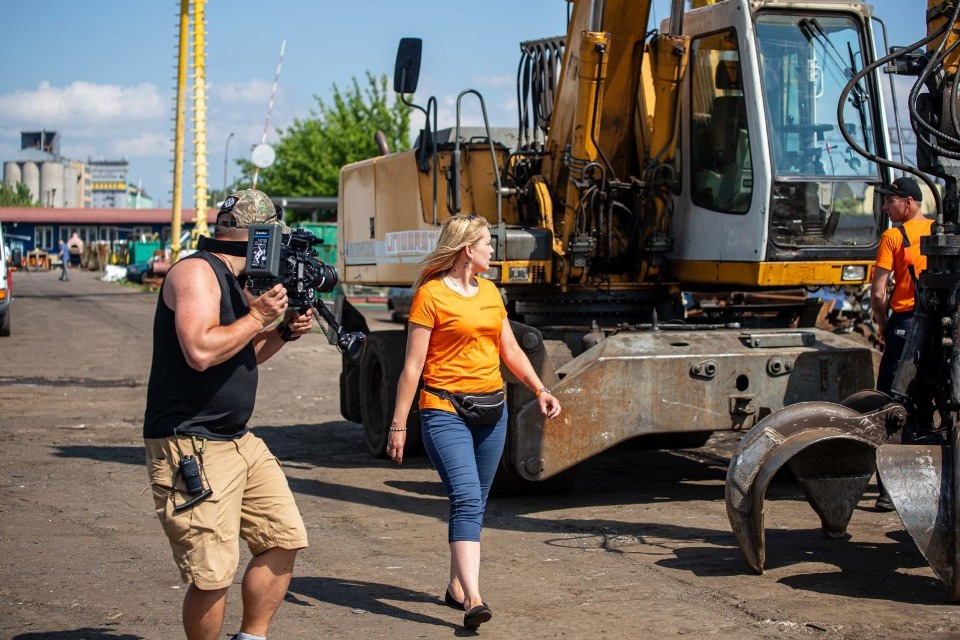 Ekipa filmowa kręci ujęcia z żółtą koparką na złomowisku; kobieta w pomarańczowej koszulce przechodzi przed kamerą.