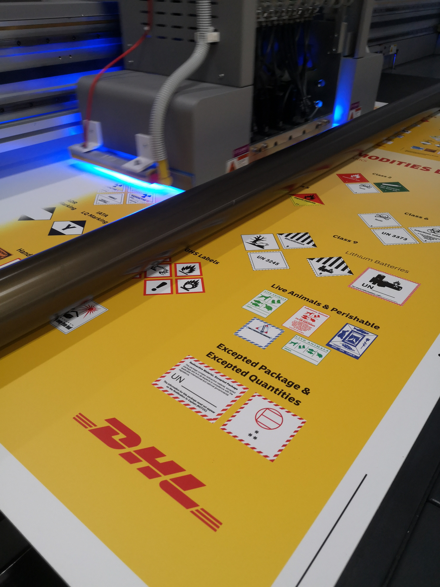 Proces druku UV na żółtym arkuszu z symbolami ostrzegawczymi i logo DHL, widoczny fragment maszyny drukarskiej z niebieskim światłem.
