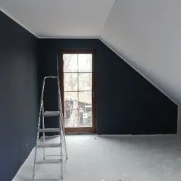 Pokój na poddaszu z pomalowaną na granatowo ścianą, białą skośną ścianą i metalową drabiną stojącą przy ścianie obok okna.