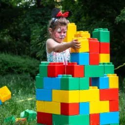 Dziewczynka z pomalowaną twarzą w tygrysa i uszami Myszki Miki układa wieżę z dużych, kolorowych plastikowych klocków na trawie.