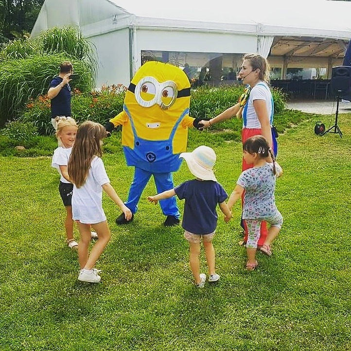 Uśmiechnięta kobieta w stroju klauna i postać w kostiumie Minionka prowadzą grupę dzieci na trawie, w tle biały namiot.