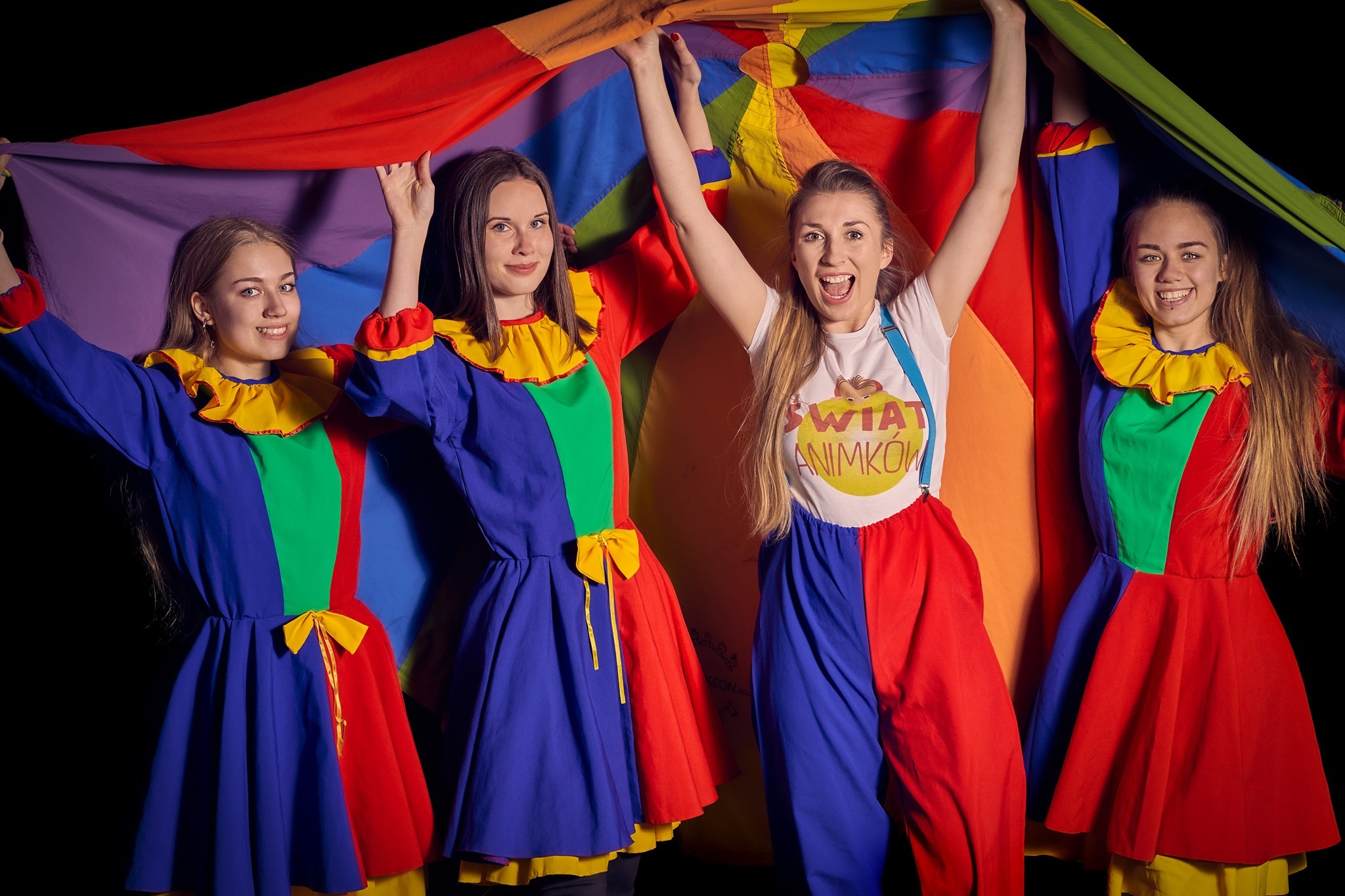 Grupa uśmiechniętych kobiet w kolorowych strojach klaunów trzymających nad głowami tęczową chustę animacyjną na czarnym tle.