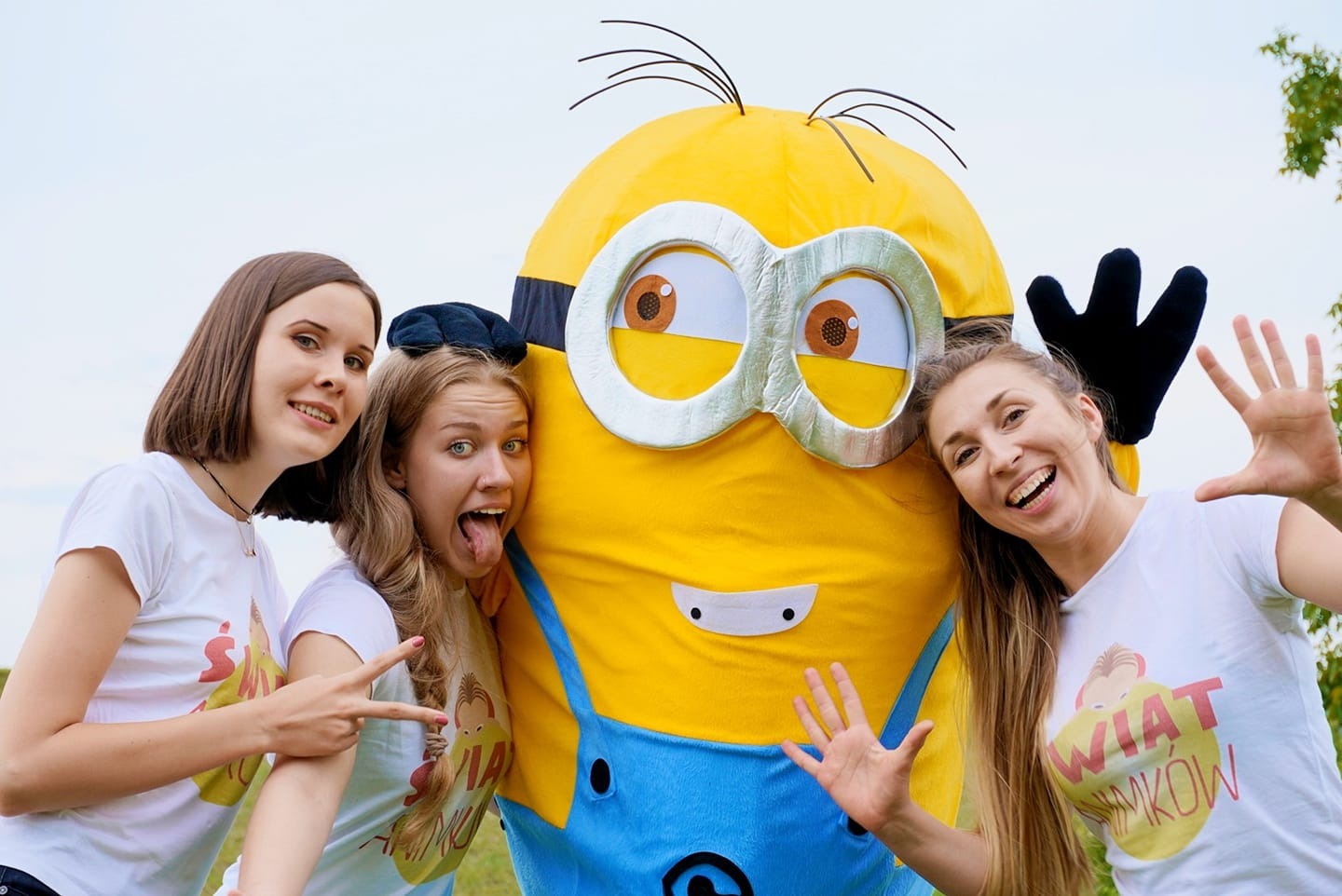 Trzy uśmiechnięte osoby pozują z dużym, żółtym kostiumem postaci z kreskówki, dwie osoby w białych koszulkach z nadrukiem, jedna pokazuje język.