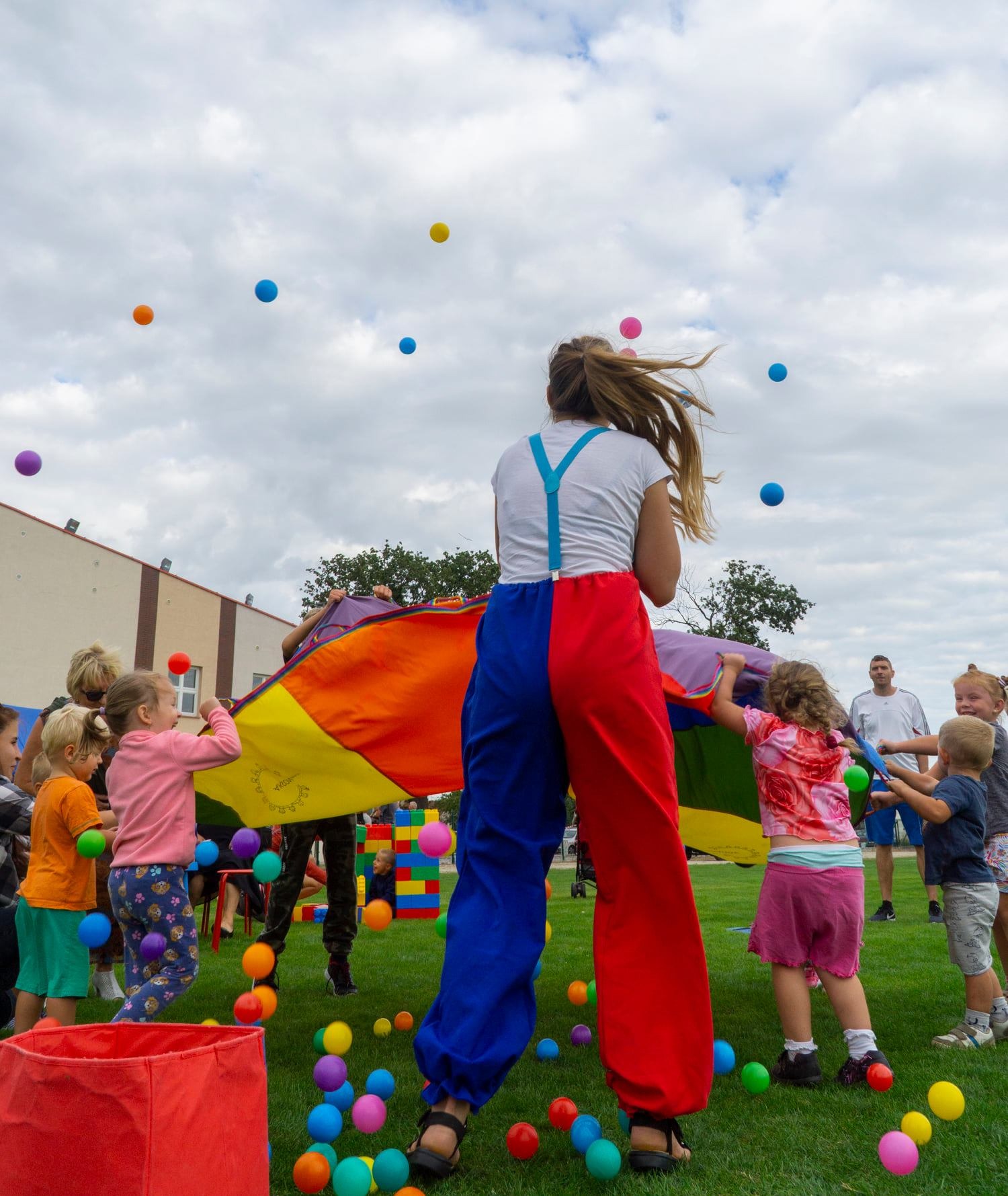 Dzieci bawiące się kolorową chustą animacyjną i unoszącymi się w powietrzu piłeczkami, prowadzone przez osobę w dwukolorowych spodniach i szelkach, na trawniku przed budynkiem.