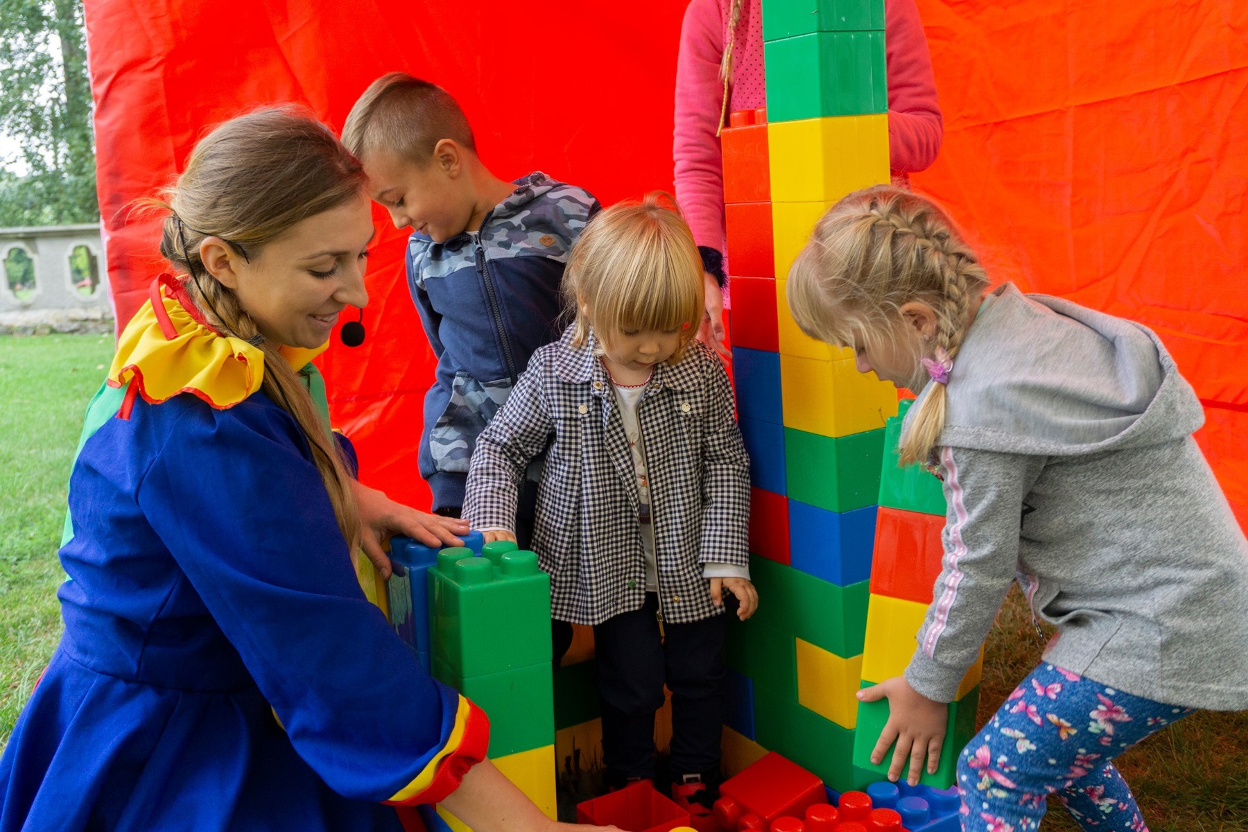 Kobieta w kostiumie klauna z mikrofonem, nadzoruje budowę wieży z dużych kolorowych klocków przez grupę dzieci na tle czerwonej tkaniny.
