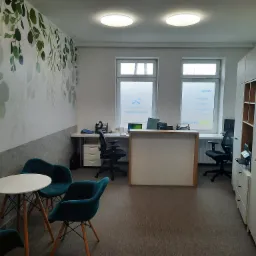 Wnętrze biura z recepcją, stolikiem kawowym i zielonymi krzesłami, dekoracyjna ściana z motywem liści, naturalne światło z okien.