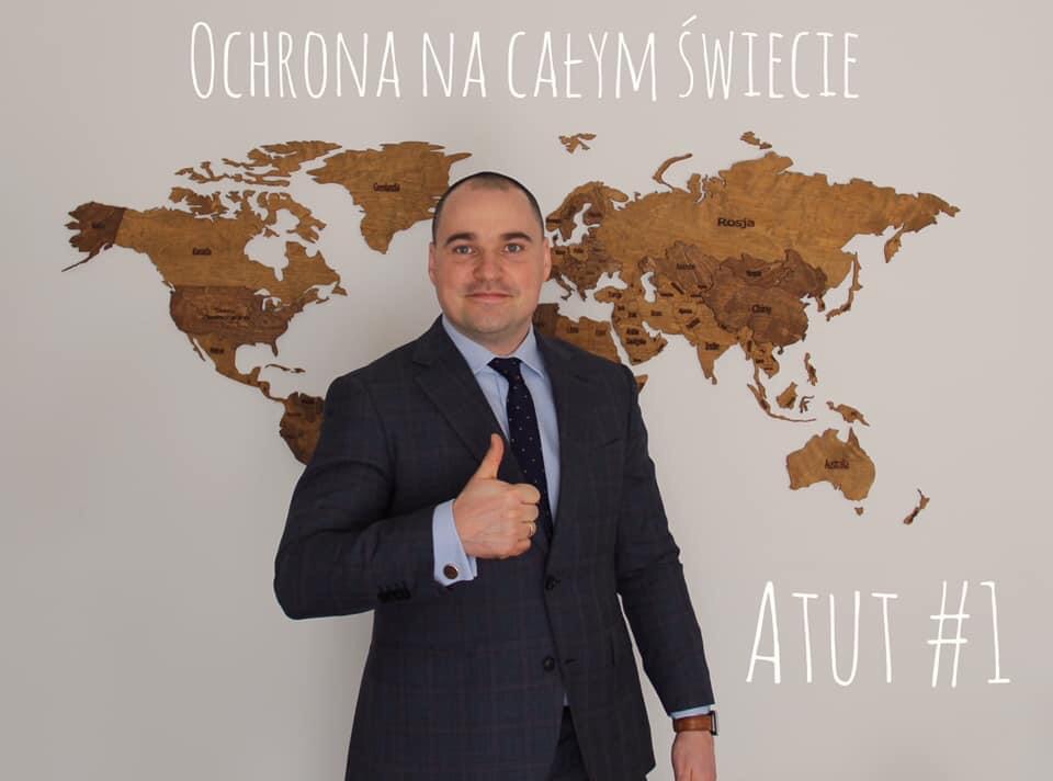 Mężczyzna w garniturze, z uniesionym kciukiem, stoi na tle drewnianej mapy świata z napisem 'Ochrona na całym świecie' u góry i 'ATUT #1' na dole.