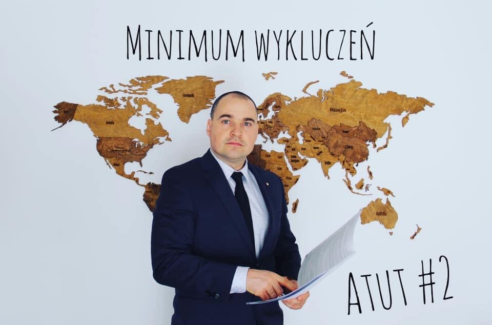 Mężczyzna w garniturze trzyma dokumenty na tle drewnianej mapy świata z napisem 'MINIMUM WYKLUCZEŃ' i 'ATUT #2'.