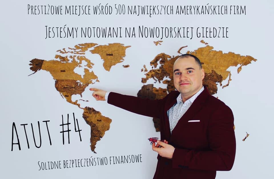 Mężczyzna w bordowej marynarce prezentuje drewnianą mapę świata z napisem 'Jesteśmy notowani na Nowojorskiej Giełdzie', wskazując palcem na Amerykę Północną, trzymając w dłoni model czerwonego...