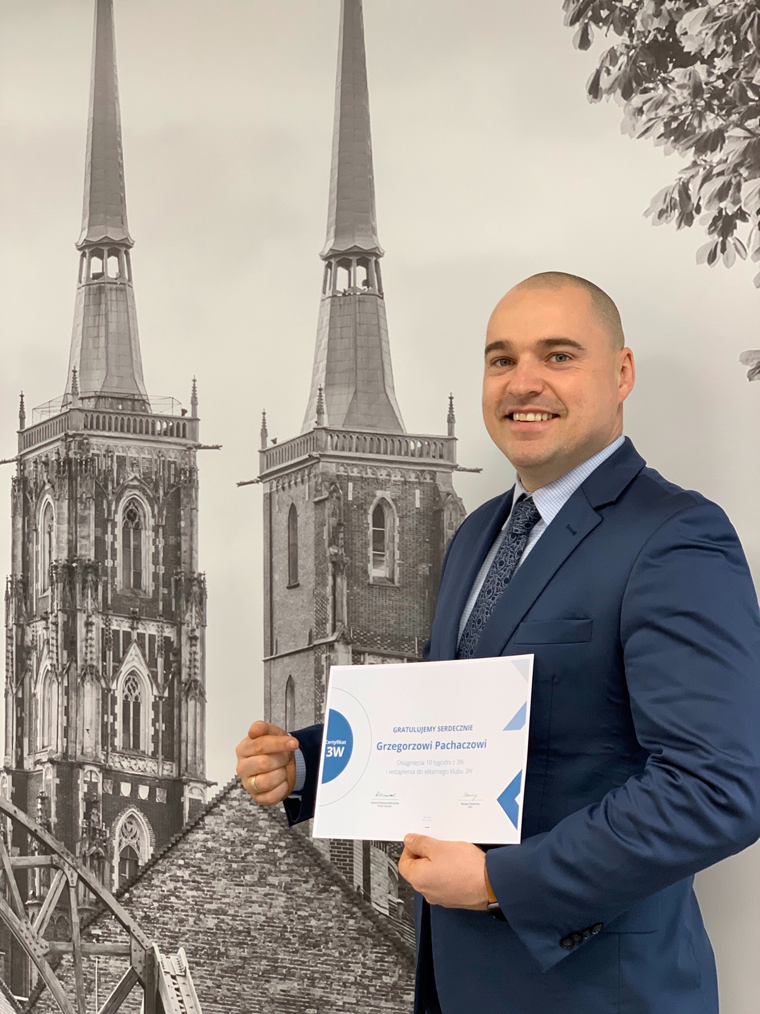 Uśmiechnięty mężczyzna w garniturze prezentuje certyfikat na tle grafiki przedstawiającej wieże kościelne i liście drzew.