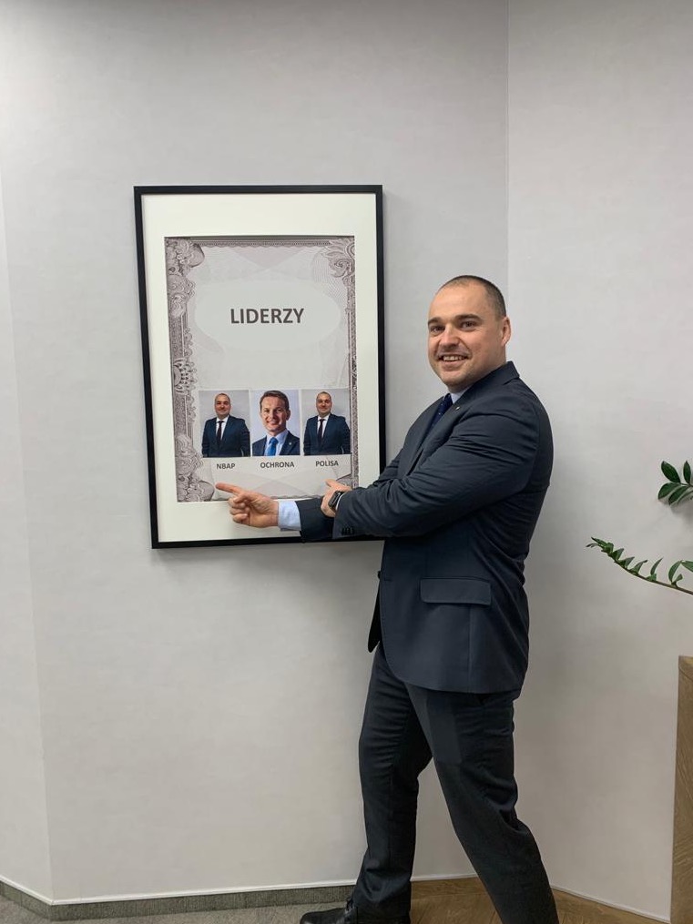 Uśmiechnięty mężczyzna w garniturze wskazuje na oprawiony plakat z napisem 'Liderzy' i trzema zdjęciami osób w garniturach, reprezentujących NBAP, Ochrona i POLISA.
