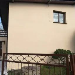 Elewacja domu po malowaniu na beżowo, widoczna drabina przy wejściu, zabezpieczona folią ścieżka i metalowe ogrodzenie.