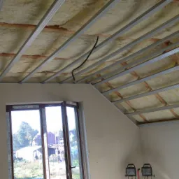 Skośny sufit poddasza w trakcie izolacji wełną mineralną, widoczne metalowe profile konstrukcyjne i fragment okna.