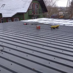 Montaż blachy na dachu w Lubaniu, widoczna zwinięta przedłużka elektryczna, narzędzia, pudełko i szczotka do śniegu, w tle ośnieżony budynek.