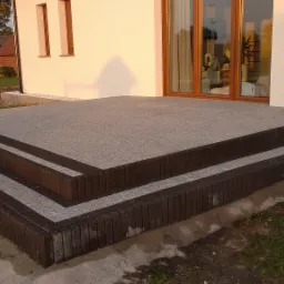 Wykończone schody wejściowe do domu z szarej kostki brukowej z ciemnobrązowym obramowaniem, widoczne okna i fragment elewacji budynku.