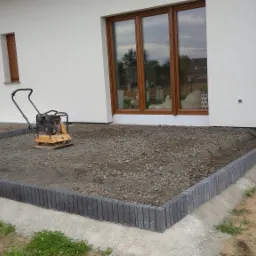 Przygotowanie podłoża pod taras: ubijarka wibrująca na żwirowej powierzchni otoczonej betonową obwódką, widok na biały budynek z brązowymi oknami.