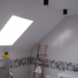 Ukośne poddasze łazienki z oknem dachowym, czarnymi reflektorami sufitowymi, dekoracyjnymi płytkami ściennymi, drewnianą drabinką z półkami, pralką i koszem na pranie.