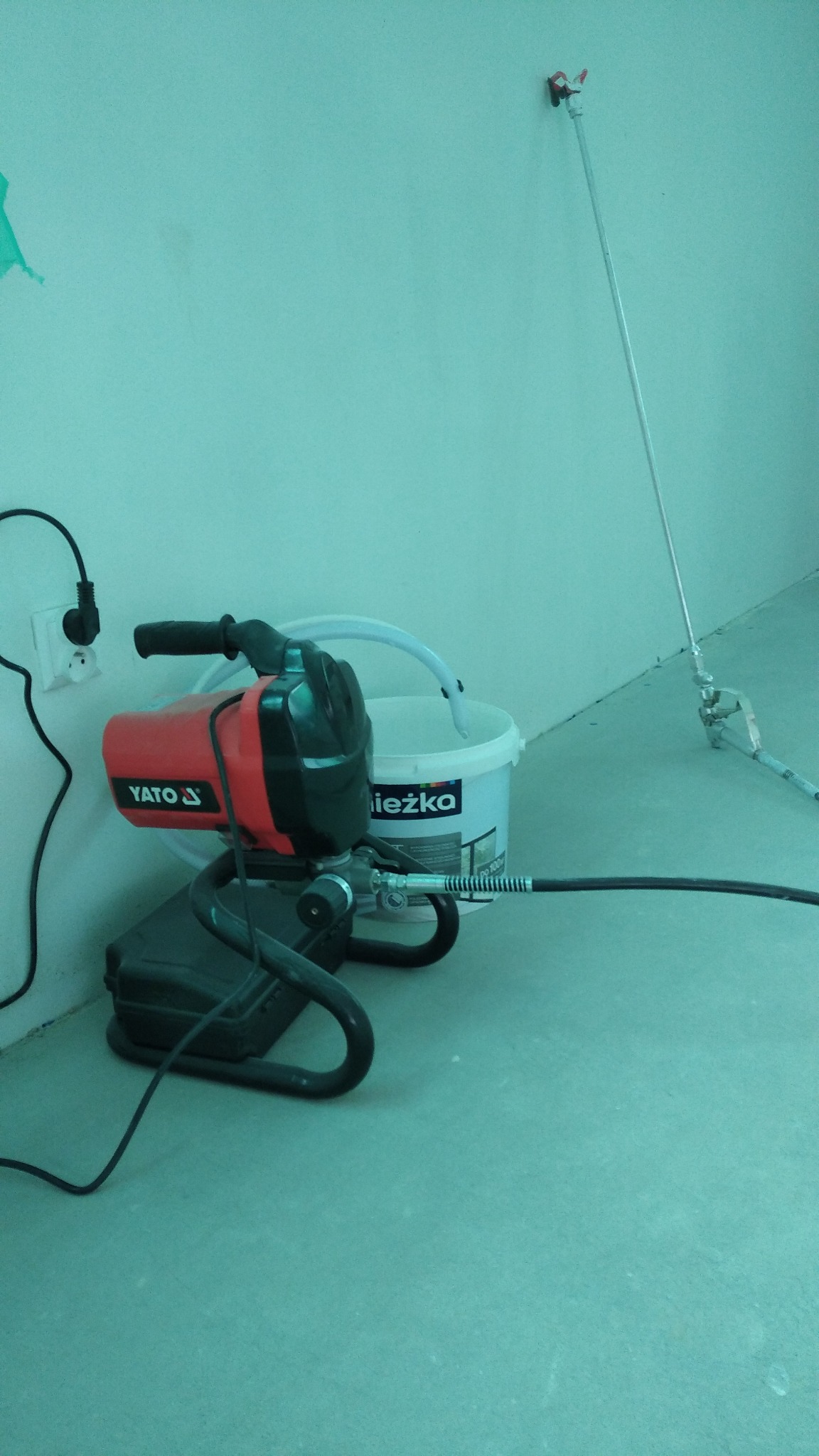 Agregat malarski YATO, białe wiadro z napisem 'Leżka' i lanca natryskowa przy ścianie w trakcie prac malarskich.