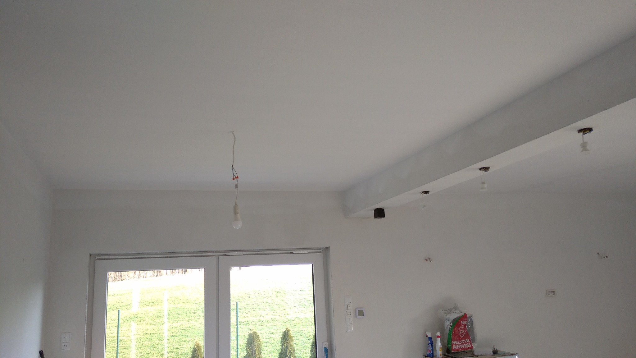 Wnętrze w trakcie wykończenia: białe ściany po szpachlowaniu, widoczne przewody elektryczne, przygotowane punkty oświetleniowe i torba z zaprawą.