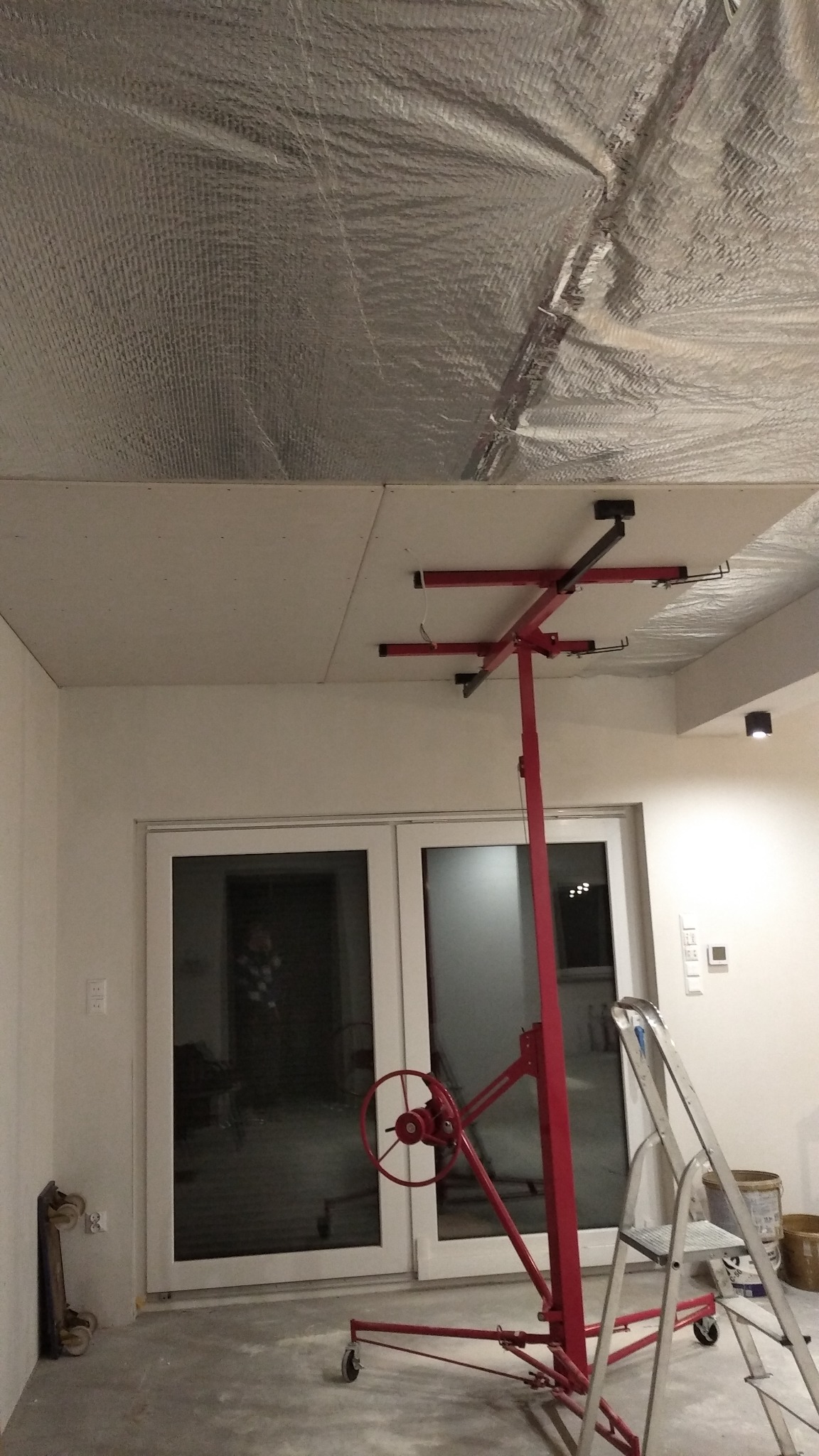 Montaż płyty karton gips na suficie przy użyciu podnośnika, widoczna izolacja termiczna, drabina aluminiowa oparta o ścianę, jasne pomieszczenie z oknami balkonowymi.