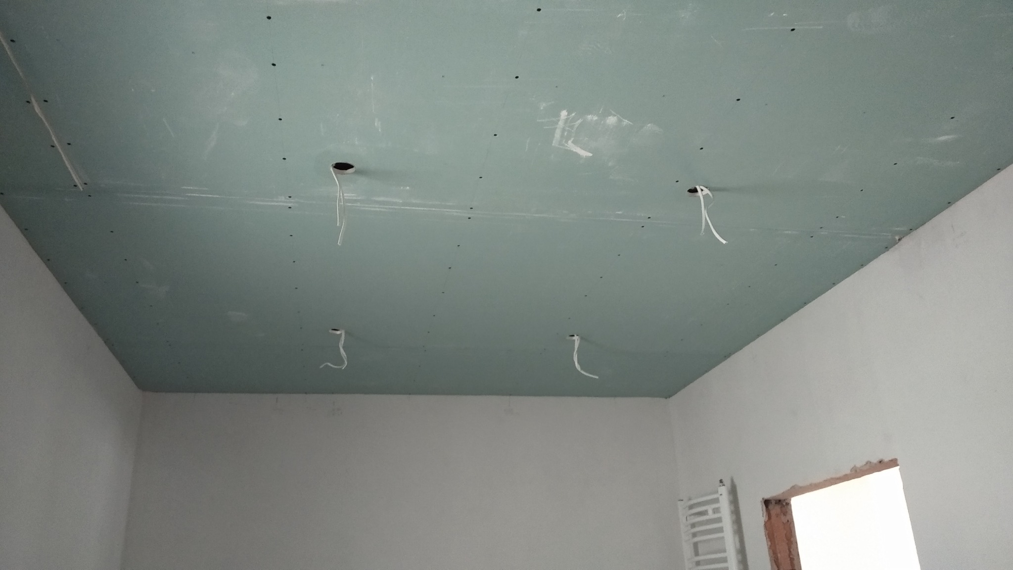 Sufit z zielonych płyt kartonowo-gipsowych, widoczne otwory na oświetlenie z wystającymi przewodami, ślady szpachlowania i wkręty, białe ściany w tle, fragment grzejnika i otworu okiennego.